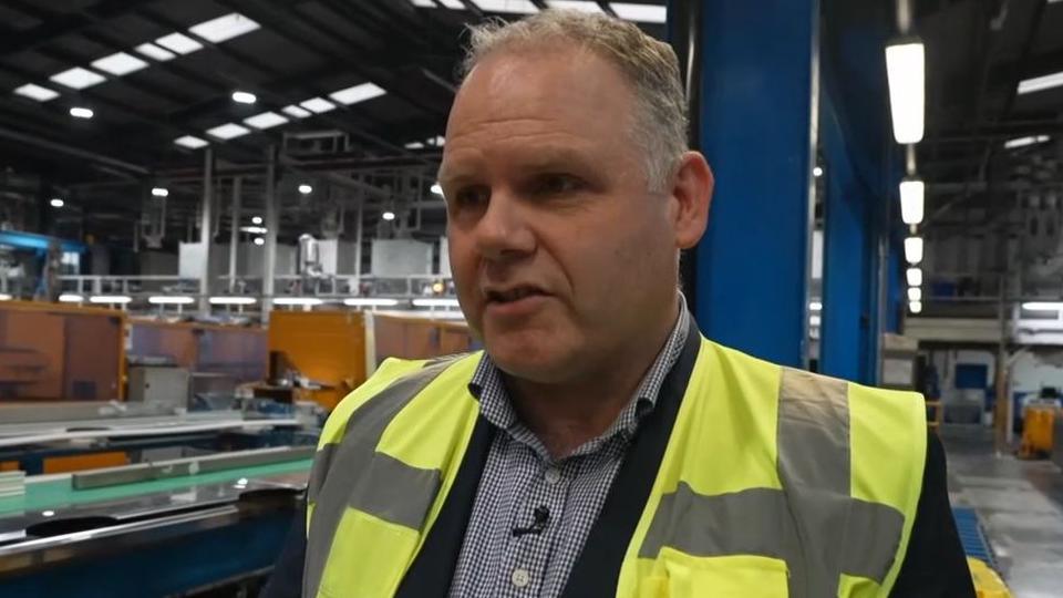 Commercial director Tim Taylor standing in a window factory in a high vis jacket and smart shirt. He has short grey hair and is wearing a smart checked shirt