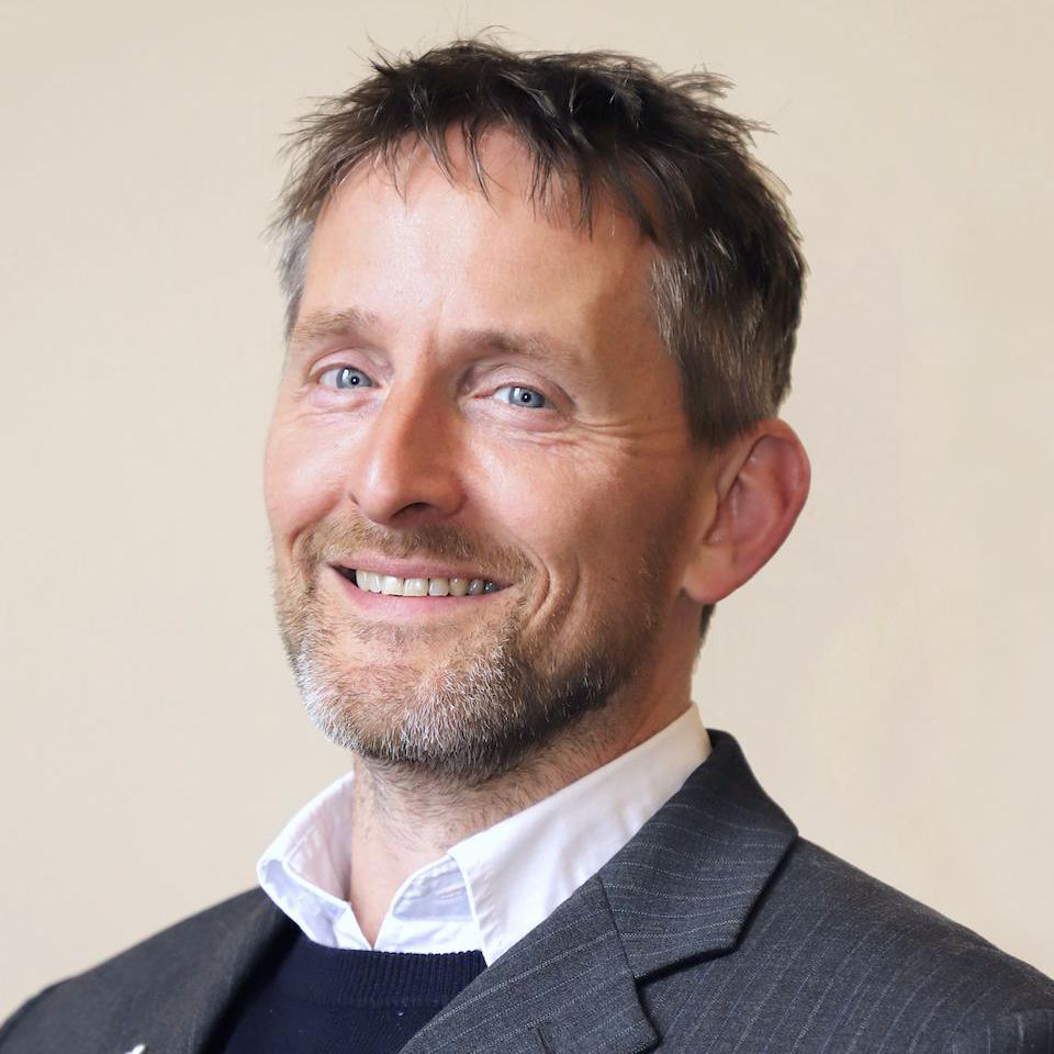 A man with greyish brown hair and a beard in a jacket with a white shirt and blue jumper underneath