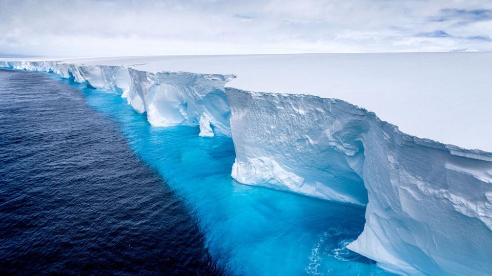 The image captures a dramatic meeting point between a towering ice shelf and the open ocean. It’s a wide, cinematic view that emphasizes both scale and isolation. The ice wall is massive and vertical, stretching far into the distance.
Its surface is textured with cracks, ridges, and natural sculpting, giving it a rugged, ancient look.
The ice appears bluish‑white, with deeper blues in the crevices where the ice is denser.The water beside the ice shelf shifts from deep navy blue to bright turquoise.
The gradient suggests varying depths and possibly meltwater mixing with the sea.
The surface is relatively calm, with gentle ripples reflecting the light.