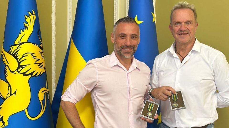 Prof Mark Hannaford and Luca Alfatti standing in front of three flags, each  holding a medal