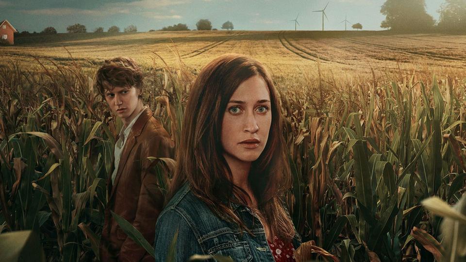 The image shows a wide rural landscape with two people standing among tall, dense corn plants in the foreground.