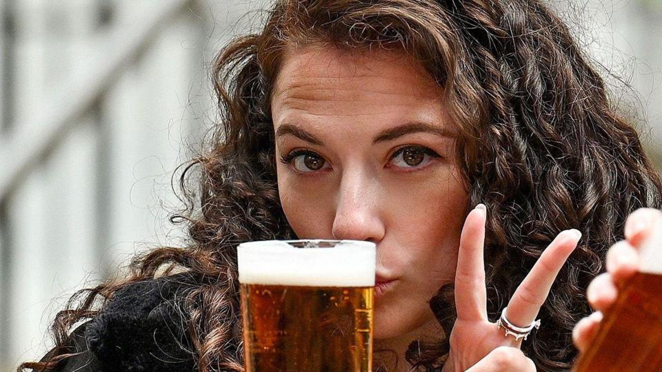 A woman and a man sitting outdoors holding pints of Lager. She is making a peace sign and he is drinking, in a casual seating area with steps and railings in the background