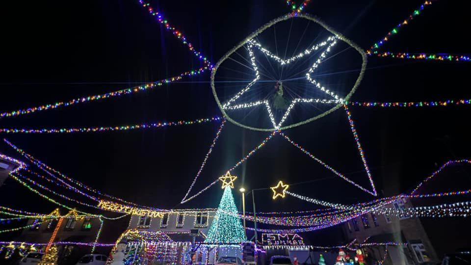 A street in Gloucestershire which is decorated with hundreds of multicoloured lights. A large Christmas tree light is in front of one house. There is a large star made out of lights which is hung up in the sky. 