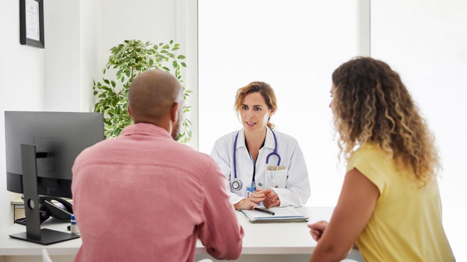 A female physician, wearing a white coast and stethoscope, discussing medical matters with patients in a doctor's office