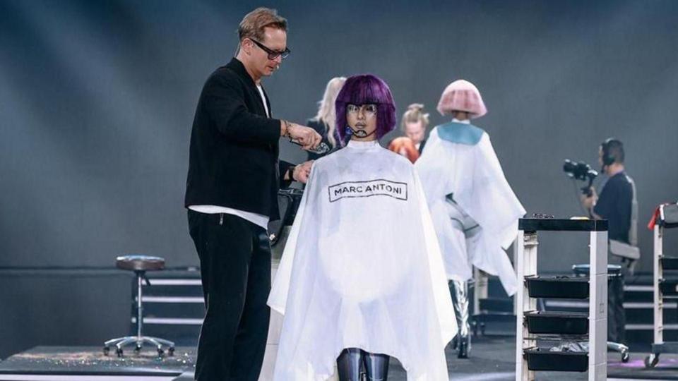 Bruno Marc Giamattei is cutting a purple wig on a mannequin. He has glasses and a black blazer on.
