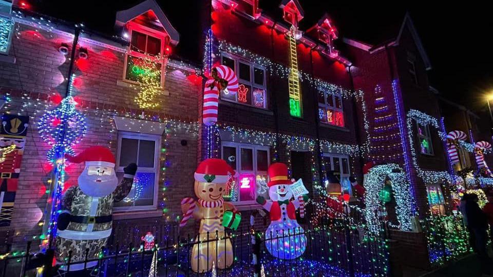 Eastbury Way, Swindon, decorated for Christmas. A few properties can be seen in the frame, all decked out with multicoloured fairy lights. There are inflatable figures in front of the houses too - a Santa, gingerbread and a snowman can be seen.  