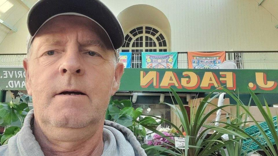 Michael Fagan in front of his plants stall. He is wearing a grey sweatshirt and black cap. There are plants and a pink bouquet behind hiim, as well as the logo of his shop, J Fagan, in yellow letters on a green background.