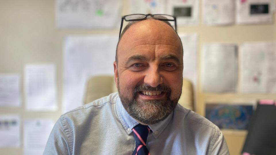 Shows a man with a beard and receding hairline with dark-rimmed glasses on the top of his head. He is wearing a blue button-down shirt with a blue and pink striped tie and is sitting in a classroom.