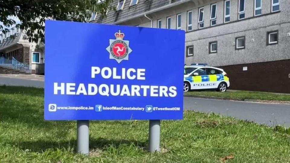 A blue sign outside a large grey building reads 'police headquarters', it also bears the Isle of Man Constabulary's emblem. A police car is parked in the background.