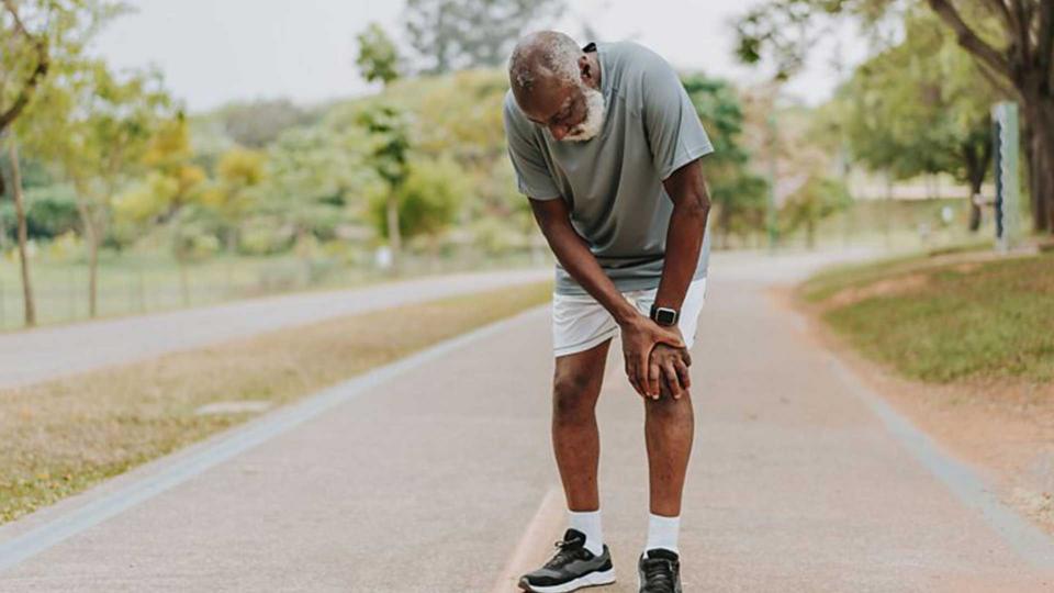 An older man wearing shorts and trainers outside bent over holding his knee.