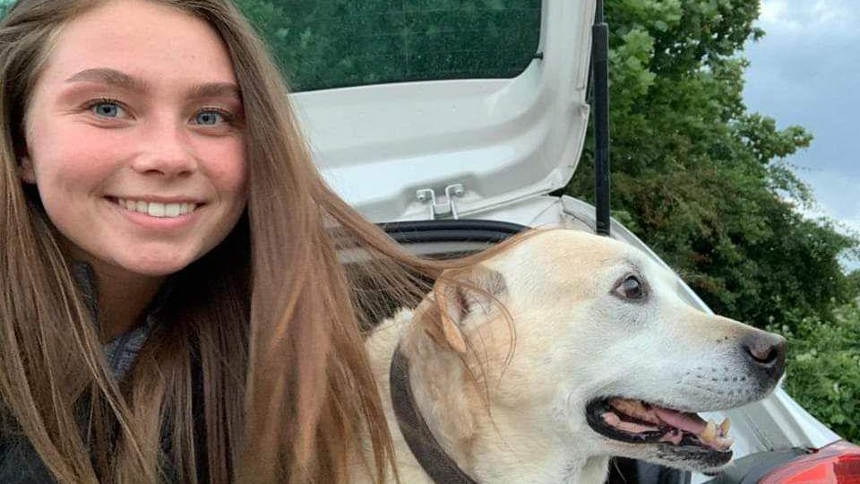 A selfie of Jaysley Beck sitting in the open boot of a white car beside a white Labrador. She has blue eyes and is smiling at the camera, with her long straight brown hair down. There are bushes and trees in the background.