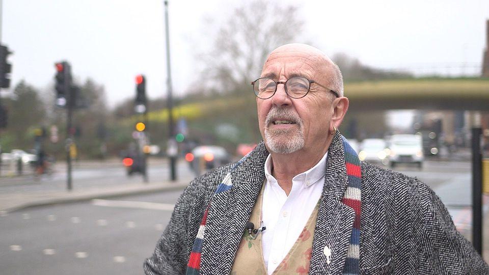 An older man, with glasses and a hoop in one ear, wearing a smart overcoat, colourful striped scarf, crisp white shirt and floral waistcoat, stands looking thoughtful in a part of East London blurred in the background