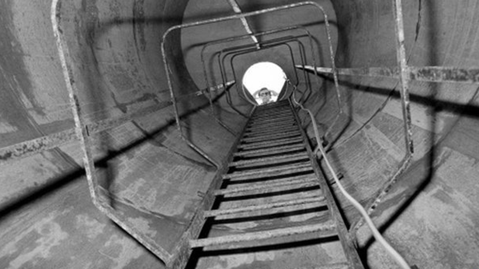 An old black and white picture from the bottom of a ladder in a drainage shaft, looking up towards the surface. A light-coloured wire is seen dangling down on the right. A person can be seen looking down from the top of the ladder.