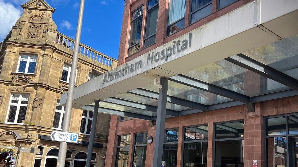 The sign above the entrance to Altrincham General Hospital on sunny day. The building below is red brick and there are two grey pillars. A sign with a blue P indicates parking and reads Regent Road pointing to the left. 
A beige stone building is next to the hospital.