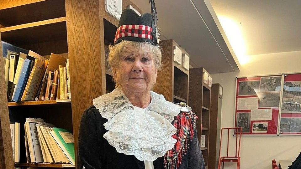 Sheila Hatcher standing in her uniform, consisting of a black velvet jacket, red tartan bagpipes, white lace collars and black hats