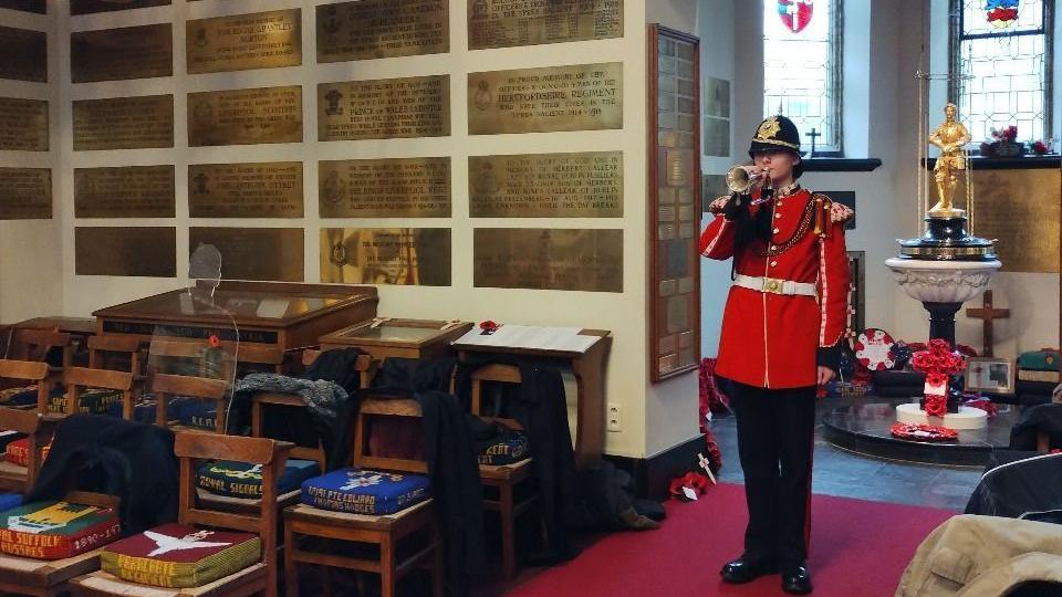 A soldier stands in a church playing a bugle. He wears a red buttoned up jacket, black hat and black trousers. There is a font behind him with a cross made of poppies at the foot, and a gold statue on top. Brass plaques line the wall.