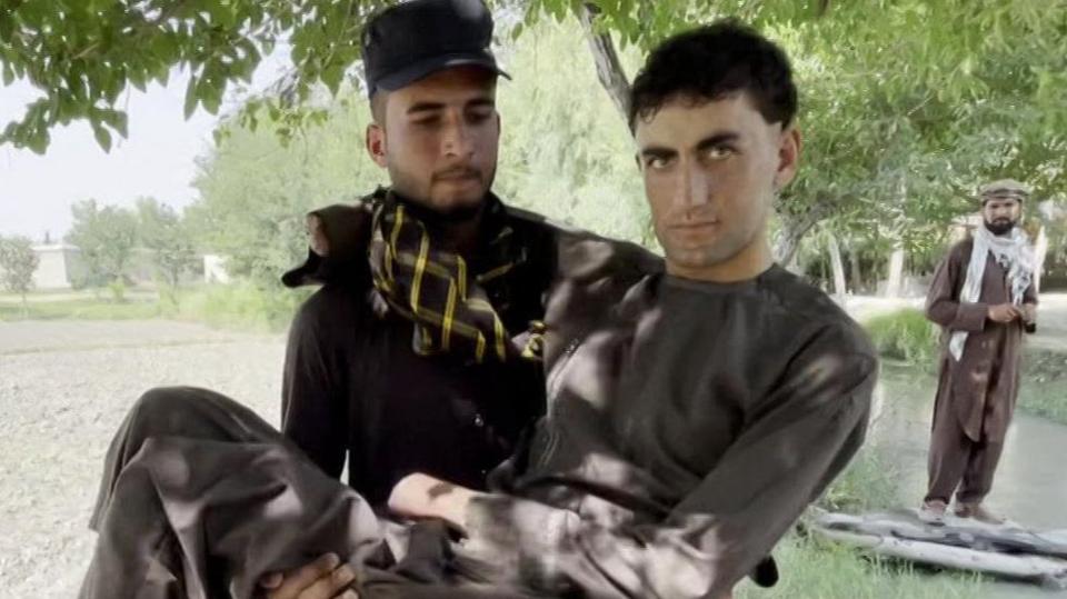 One of the 12 migrants who spoke to BBC Afghan Forensic, quadruple amputee Shahsawar, 21, is carried by another man, on a red-and-blue striped rug, while a third looks on, in a tree-lined Afghan landscape. All wear brown salwar-kameez, the two unnamed, bearded, men hats and patterned scarves.