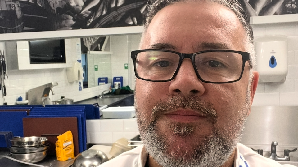Mark Petrou has short black hair with grey flecks and matching beard and moustache. He is wearing black framed glasses. He is behind the counter of a fish and chip shop.