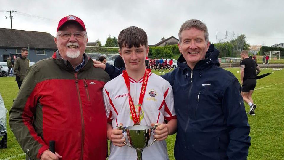 An elderly man in a red north face zip up and red cap and glasses. He has a white goatie and is smiling at the camera. Beside him is a boy with a long brown fringe in a white football jersey holding a trophy with a red medal around his neck. Beside him is a man in a navy waterproof coat with grey short hair, smiling. Behind them is a football team warming up.