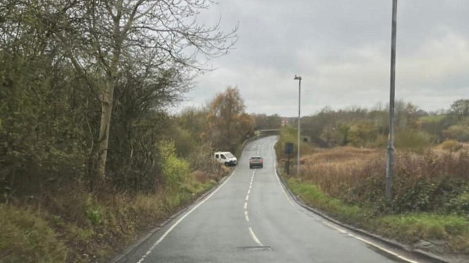 A view from Bullcote Road in Oldham, with a white van seen pulling out by a grey car in the distance. Streetlights can be seen either side of the road, which does not have a pavement, and runs through a rural setting. 