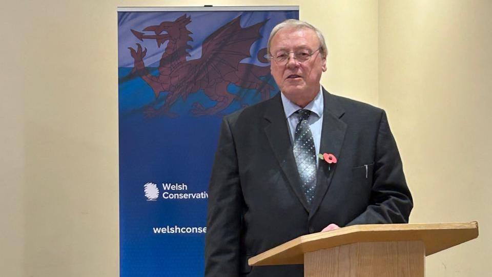 Bernard Gentry wearing a black suit, with a poppy attached, speaking in front of a Welsh Conservative banner.