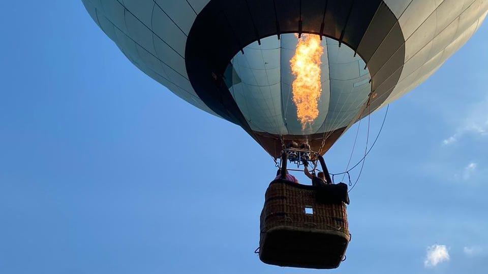 The underside of a hot air balloon with a bright orange flame as its burner is fired. In the background the sky is almost entirely blue, with some small white clouds
