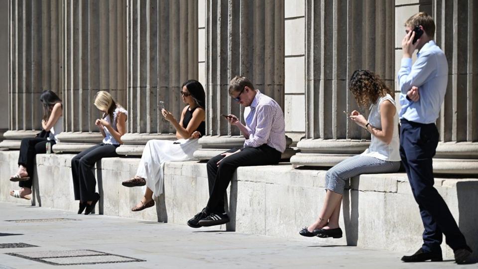 Five people sit along a low wall in the sunshine looking at their mobile phones, while a man stands at the right-hand side speaking to someone on the phone