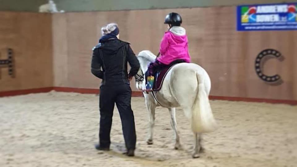 A child in a pink jacket rides on a white pony, an adult wearing black trousers and a black jacket walks next to them.