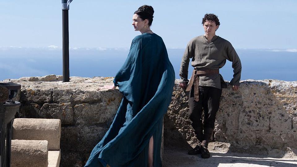 Anne Hathaway as Penelope (Odysseus's wife) in centre of image in blue gown looking to the side and Tom Holland as their son Telemachus leaning on wall in khaki top and dark trousers.
Blue skies behind
