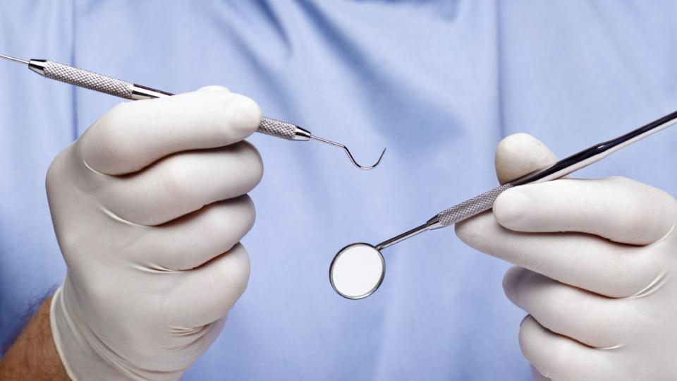 A dentist wearing a blue gown white gloves holding dental instruments