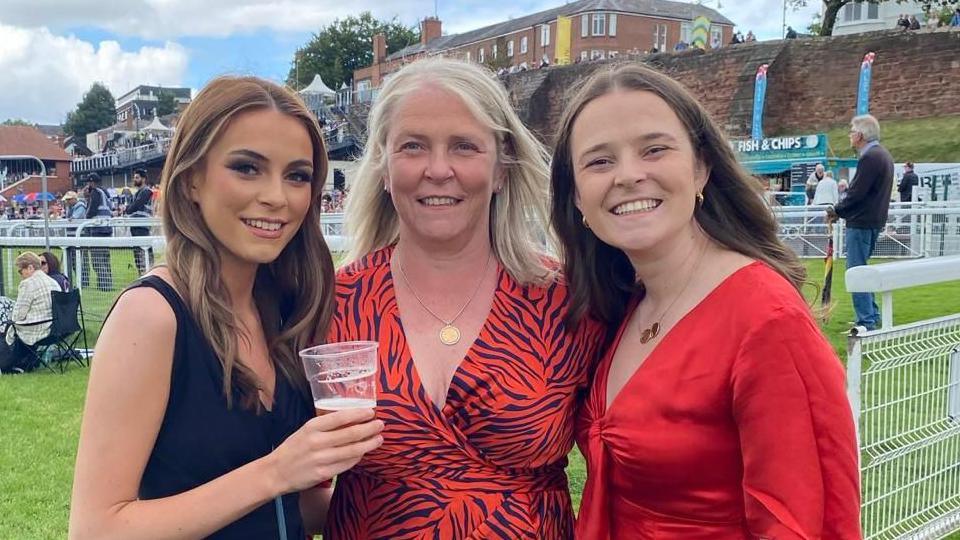 Three women stood side by side, all looking into the camera and smiling. In the middle is Lisa, wearing a red and black tiger-print dress, and either side of her are her daughters, wearing black and red dresses. They're stood outside at what looks like a racecourse.