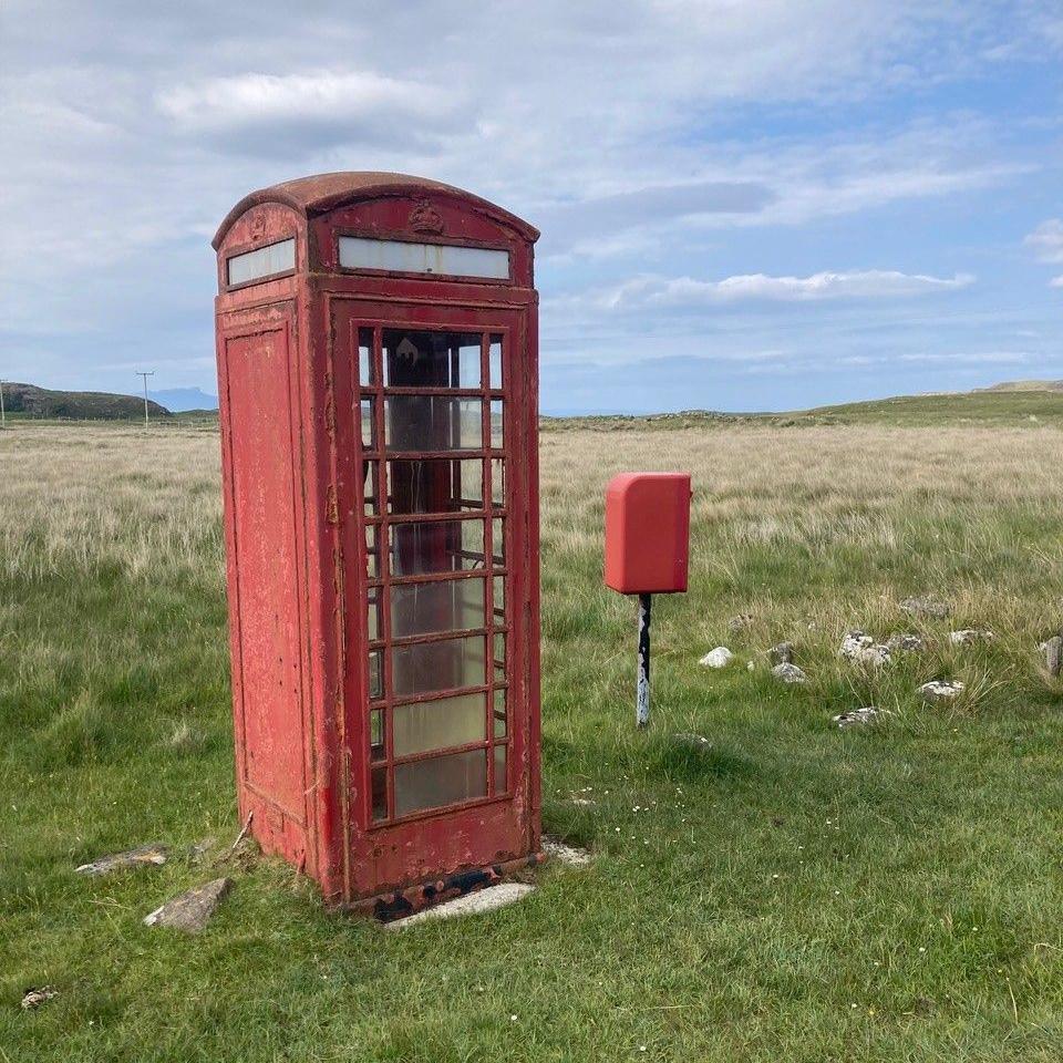 A red phonebox in a grassy field. It has been damaged and has bits of paint coming off it. A small red postbox facing away from the camera is to its right. They are both on a grass patch with longer grass behind it.
