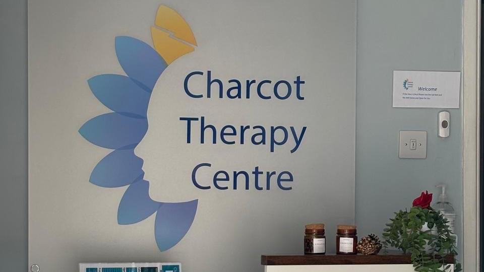 The foyer area of Charcot, there is a large white sign reading 'Charcot Therapy Centre'. On the table there are small brown jars, decorated pine cones and a green plant with a red rose.