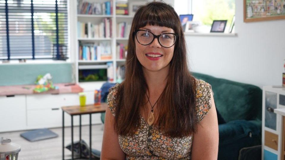 Heidi smiles at the camera. She has long dark hair with a thick fringe and large black glasses. She wears a khaki green, yellow and white summer dress and a wooden necklace. Behind her is a green sofa and white bookshelves.