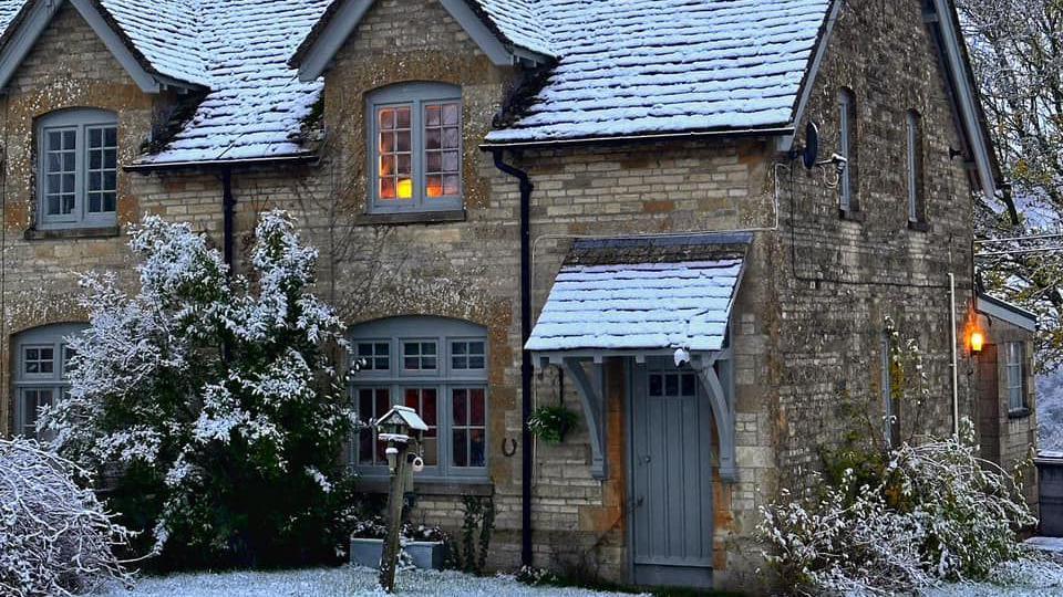 A cosy image showing a country cottage with orange lights on in the sash windows and snow settled on the front door awning and trees, grass and bushed outside. A thick sheet of snow has also covered the tiles on the roof.