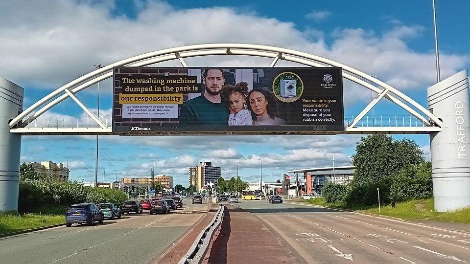 An advertising board on the side of a foot bridge over a dual carriageway in Trafford. It shows a family standing outside their house with the words 'The washing machine dumped in the park is our responsibility'