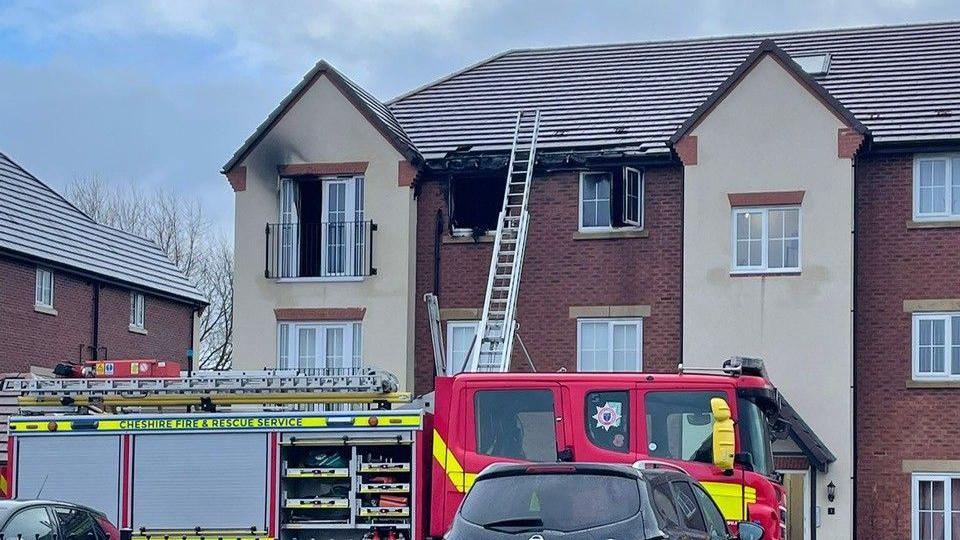 A fire engine in front of a flat with three burnt out windows. A ladder is leaning against the front of the property and reaches all the way up to the roof.