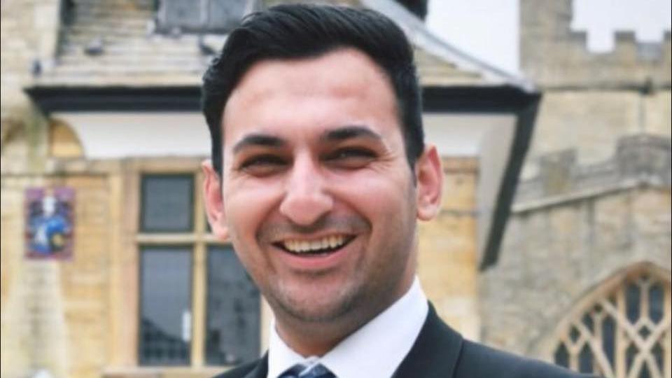 Nabil Ilahi smiles and looks directly at the camera as he is photographed in suit and tie and standing in front of a church