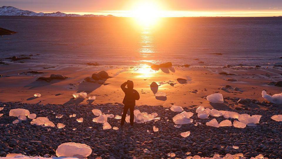 A person stands on a beach at sunset among chunks of ice washed up on the shore in Nuuk, Greenland.