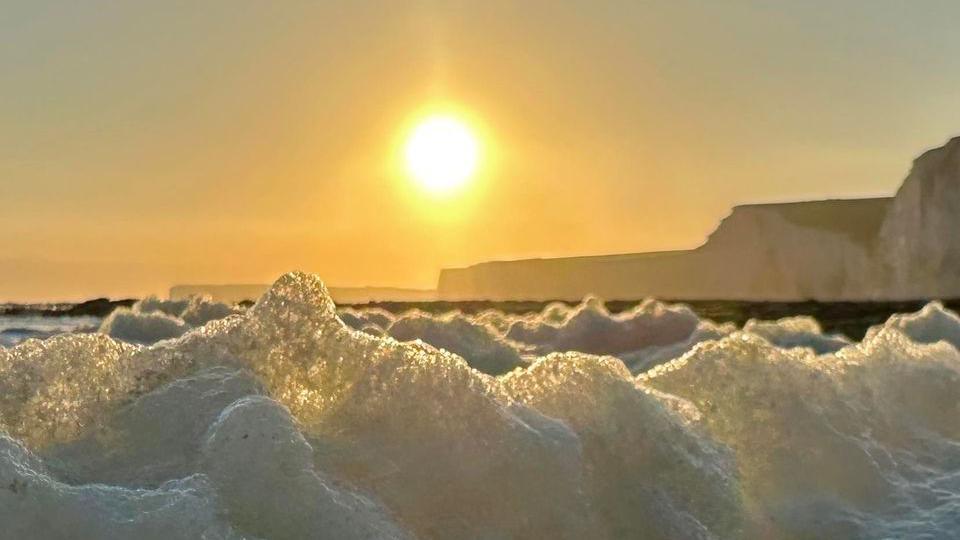 A close of sea foam with the sun and cliffs looming in the background 