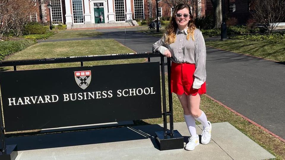 Amy Donohoe wearing a striped shirt with red skirt. She has sunglasses on. She is leaning on a sign that says Harvard Business School.