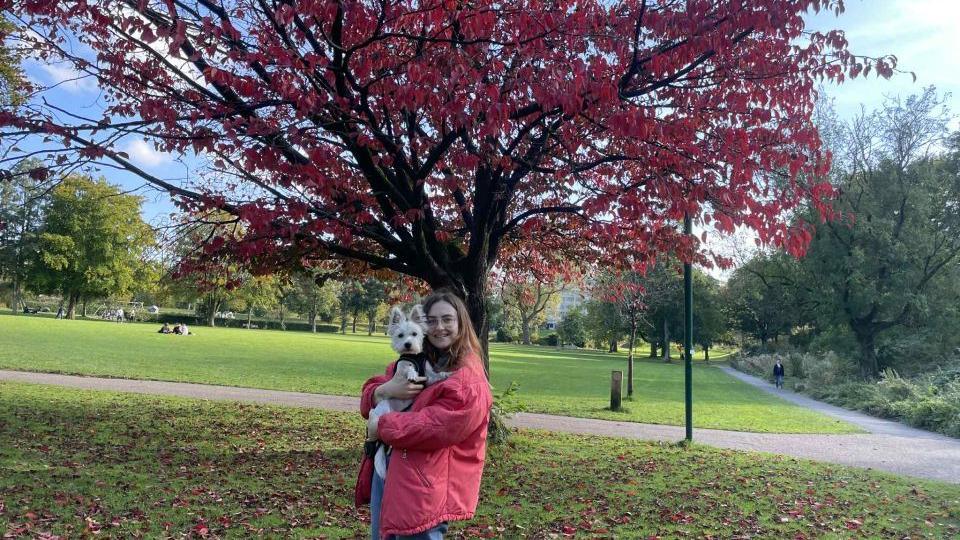 Chloe Price, who is wearing a red jacket, is holding a dog. There is a tree in a red autumn colour behind her.