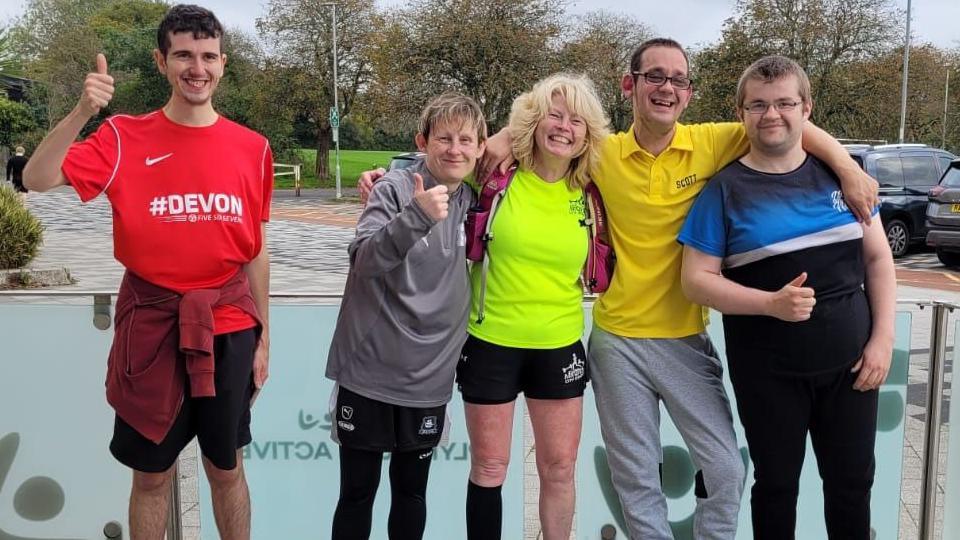 A group of five people smiling with their arms around each other's shoulders giving thumbs up signs while wearing running clothing and trainers.