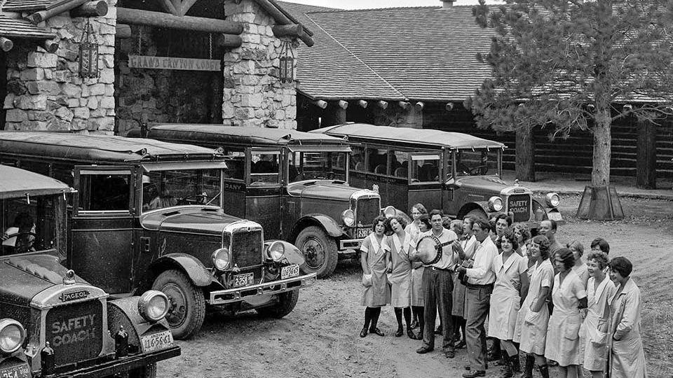 A black-and-white photo of old-fashioned buses in front of a stone building, with a line of women in cleaning uniforms and two men playing stringed instruments