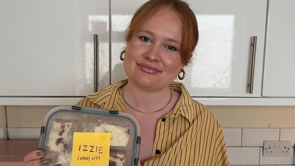 A woman holding a lunch box that has a sticker on it saying "Izzie (Hands off)"