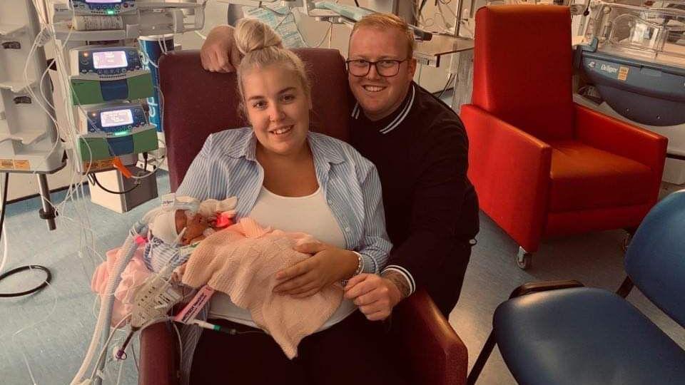Chloe and Kyle pose for a picture with a new-born Ella in a hospital room. Chloe is sitting on a red hospital chair holding the baby as Kyle crouches to pose.