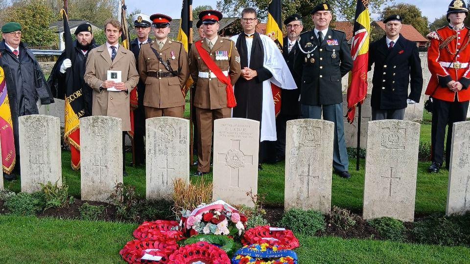 Seven identically shaped headstones in a row, facing forwards, engraved with a cross and words. Wreaths of poppies are laid in front of the newest grave, and behind the graves stand a group of people. One is a vicar wearing white robes and a black cassock, others are wearing brown or black miltary uniform and holding flags. One man appears to be a family member, standing holding an order of service.