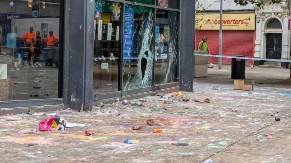 Cosmetics of various colours are strewn across the pavement in front of a Lush store, which has a smashed window