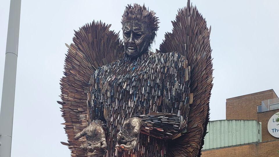 The Knife Angel, a 27 ft (8m) sculpture made from 100,000 blades. It is a male figure, looking down, holding out its hands palm up, with wings behind it.