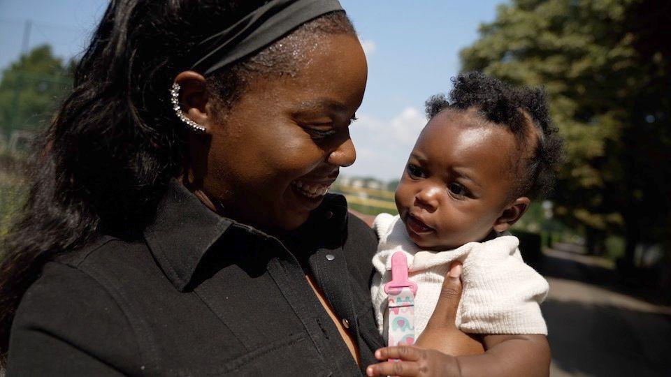 A woman dressed in black smiles at her 6-month-old baby who she carries in her arms. They are in a sunny park.